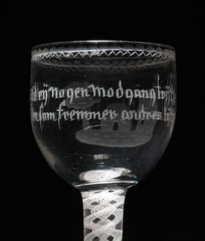  An ornate glass goblet with a spherical body and a latticed stem, etched with decorative patterns and gothic script, against a dark background.