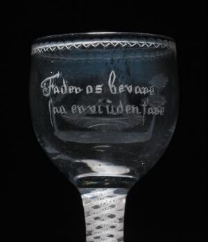  Antique glass goblet with diamond point engraving and etched text around the rim, set against a solid black background, highlighting the decorative features and contours of the glass with a play of light and reflection.