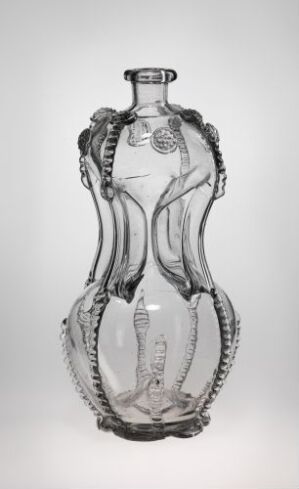  A transparent glass bottle with an hourglass shape and decorative swirled patterns, created by artist Gjøvik Verk. The piece features twisted ridges alongside the body and stylized circular details at the center, set against a simple light background.