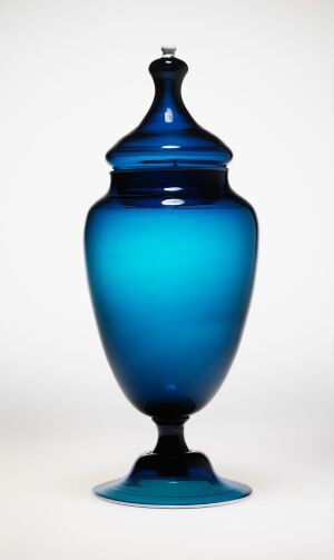  A glass vessel with an ombre effect transitions from cobalt blue at the base and lid to sky blue in the center, standing against a white background.