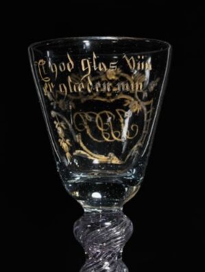  An historical glass with tulip-shaped cup featuring decorative golden calligraphy and floral patterns, set against a black background. The stem exhibits a twisted design that adds texture.