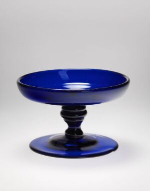  An elegant cobalt blue glassware piece featuring a shallow bowl on a tiered pedestal base, with reflective properties highlighting the depth and clarity of the blue color throughout. Artistname and title: unknown