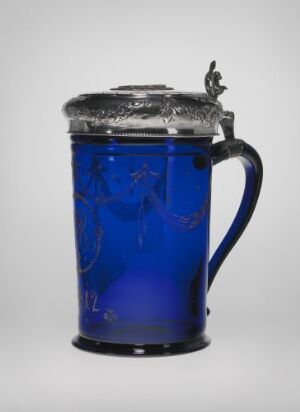  A traditional cobalt blue glass beer stein with etched designs and a pewter lid featuring a small decorative figurine on top, set against a simple gray background.