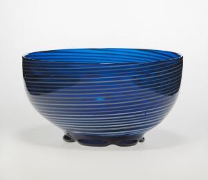  A modern bowl with a striped pattern in various shades of blue, ranging from deep navy to bright electric blue, against a light-colored background, suggesting a glass or glossy ceramic material. The bowl appears to taper toward a raised base, possibly resting on small feet.