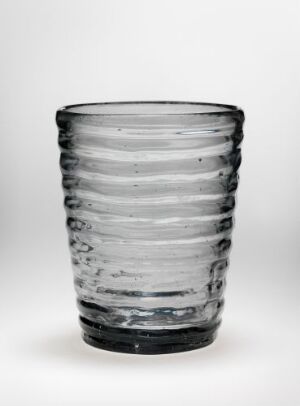  A clear cylindrical glass tumbler with horizontal ridges set against a light background, exemplifying handcrafted glassware with a textured design.
