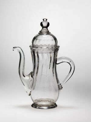  A transparent glass coffee pot with an elegant cylindrical shape, a curved handle, and an ornate spout set against a light background. It has a removable lid with a decorative knob and intricate etched details around the neck and lid.