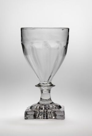  A clear glass goblet with an ornate base and a smooth stem, standing against a plain, light background.