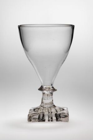  An elegant transparent glass with a conical shape atop a detailed stem and a square base with etched figures, set against a light-grey background. Artist name and title are unknown.