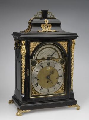  An antique, black and gold ornate clock with Roman numerals on a lighter colored face, decorative gold accents and a glass front panel, supported by four small gold feet.