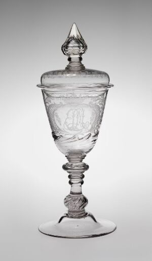  An intricate clear glass goblet with a lid, displaying a monogrammed emblem and ornate designs, stands against a neutral gray background.