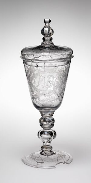  A Nøstetangen glassverk engraved glass vessel with a conical body adorned with elaborate engravings, a structured stem with a layered design, and a matching lid with a spherical finial. The piece stands against a white background, showcasing its transparent and reflective qualities.