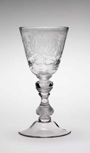  A transparent glass goblet with intricate engravings on the bowl and a twisted stem design rests against a light gray backdrop, displaying its delicate artistry and reflecting the gentle light.