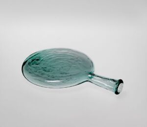  A vintage-looking oval-shaped glass bottle with a green tint lying on its side against a light gray background. The glass has a hand-blown texture with an irregular surface.