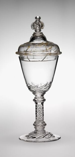  A tall and intricately crafted clear glass goblet with a lid on a white background. It features a decorated bowl with etched details, an ornate stem with bead-like designs, and a solid, flat base. The artist's name and title are unknown.