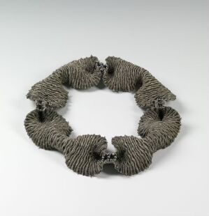  A necklace titled "Sjøanemone 1" by Ingrid Larssen, resembling a gray sea anemone made of silk, with detailed embroidery creating a textured, circular pattern.