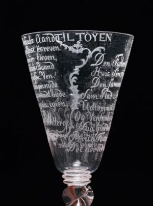  An intricately etched opaque glass with flared rim and faceted stem, featuring bold script and decorative imagery, set against a dark background. The glass has a historical appearance, with a partially visible base that may include subtle coloration.