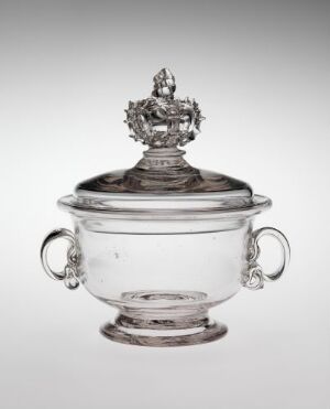  A clear glass soup tureen with ornate handles and a decorative finial on the lid, set against a light greyish-white background.