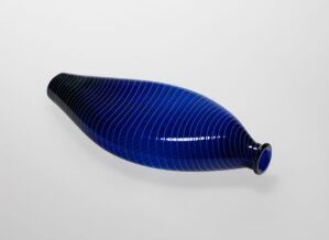  A blue glass object with a fish-like, elongated form and raised parallel lines on a light background, evoking a sense of marine life.