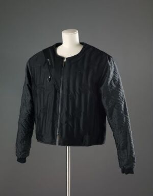  Men's black polyester jacket by L-Fashion Group OY/Rukka, with quilted sleeves and a full-length zipper, displayed on a mannequin torso against a neutral background.