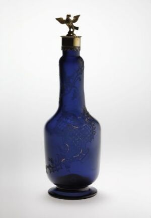  A slender cobalt blue bottle with a textured surface and an ornate metal stopper featuring a small figure reminiscent of a bird with outstretched wings, set against a neutral background.