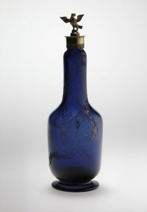  A vibrant cobalt blue glass bottle with a brass or gold-colored fixture and decorative element resembling two tiny birds or fleur-de-lis on the neck, set against a light background.