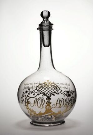  An ornate glass bottle with a pear-shaped body and a spherical stopper, showcasing a gold-colored, decorative monogram and scrollwork design on its surface, set against a plain light gray background.