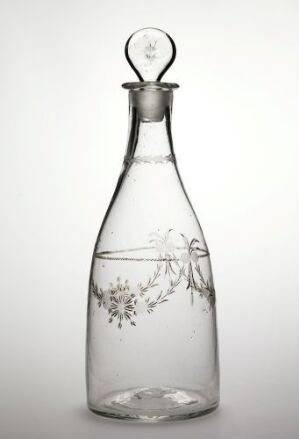  A clear glass bottle with an etched vine design near the base and an elegantly crafted stopper stands against a light grey background.