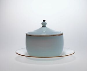  A pale blue ceramic sugar bowl with a domed lid featuring a cobalt blue knob, sitting on a matching plate with a gold rim, against a light gray background.