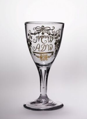  A clear glass wine goblet with black and gold decorative patterns and monograms on its upper half, positioned against a graduated white to gray background. Artist and title unknown.