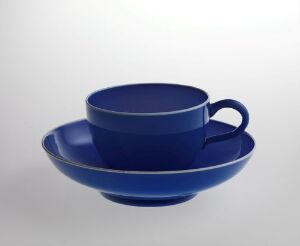  A cobalt blue ceramic tea cup with a matching saucer, set against a soft gray gradient background. The tea cup has a glossy finish and a C-shaped handle on the side.