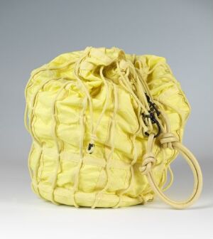  "Parachute Shoulder Bag" by Helmut Lang, a pale yellow cotton shoulder bag with diagonal grid pattern of cotton bands and leather handle, displaying a ruched, textured aesthetic against a gray background.