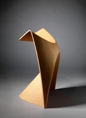  A modern sculptural furniture piece by Illums Bolighus, made from laminated and molded oak wood with a honeyed brown color. The object has an abstract S-shaped form with visible wood grain, set against a gradient gray background.