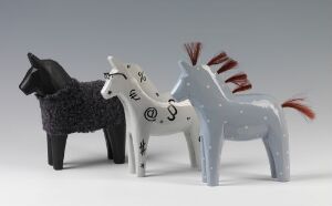  Three wooden Dalahest horses, showcasing Swedish folk art by Katarina Brieditis. The horse on the left is black with white speckled patterns, the center is white with whimsical black markings, and the right is a gray with red accents and a heart design.