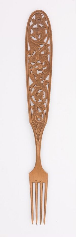  A tall, slender wooden object with a handle that tapers into an oval head elaborately carved with lacy, openwork patterns, and a fork-like base with several tines, set against an off-white background. Artistname and title are unknown.