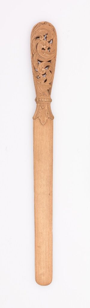  An elongated, narrow wooden spoon with intricate faces and patterns carved at its bulbous head, set against a light background, highlighting the natural beige and tan colors of the wood.