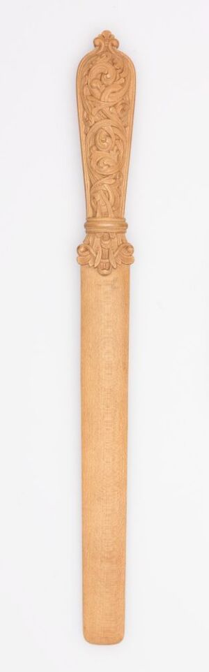  A slender wooden ceremonial staff or scepter with intricate carvings at the top, set against an off-white background. The staff is warm honey-tan in color with visible wood grain and a detail-rich, crown-like top feature. Artist name and title unknown.