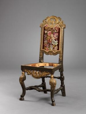  An ornate wooden chair with gold accents and a tall, tapestry-upholstered backrest featuring a red-dominated pictorial scene, set against a neutral gray background. The detailed wood carvings include scroll, foliage, and possibly floral or fruit motifs, with clawed feet and ornate cross supports. Artist name and title are unknown.