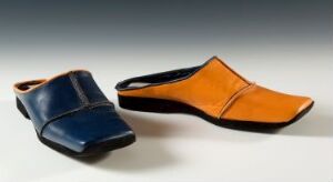  Two stylish shoes on a gradient background; one is a glossy navy blue clog on the left, and the other is an angular burnt orange slip-on shoe with a black sole on the right.