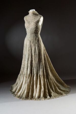  A designer gown by Hege Mari Sørum in neutral shades of creamy ivory to earth-toned beiges made from lustrous machine-woven silke, with a fitted bodice and a wide, elegantly draped skirt. The dress features subtle metallic threads and is displayed against a dark background.