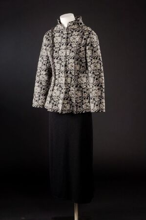  A mannequin dressed in an elegantly tailored floral jacket in white with shades of gray and black, paired with a simple black mid-calf length skirt, against a dark background.