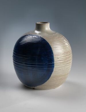  A two-toned ceramic vase with a bulbous body and a narrow neck. The left side has a glossy cobalt blue glaze with raised horizontal ridges, while the right side features a cream or off-white glaze with fine vertical etchings. The vase is set against a neutral grey background. Artist and title unknown.