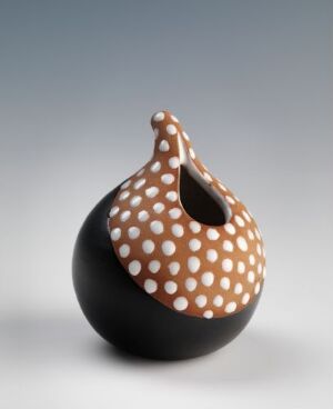  A ceramic vessel with a rounded base and an asymmetric looping opening at the top, featuring a black matte finish on the lower half and glossy white with black polka dots on the upper half against a soft gradient background. The artist name and title are unknown.