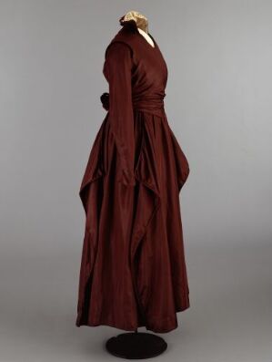  A historical burgundy dress with long sleeves and a full skirt displayed on a mannequin against a light gray background, embodying late Victorian or Edwardian era fashion elegance. Artist name and title are unknown.