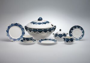  Ceramic tableware set designed by Andreas Schneider, including a large soup tureen with lid, a dinner plate, a smaller plate, a cup with saucer, and an extra saucer, all in white with hand-painted blue jagged patterns around the edges, presented against a soft grey gradient background.