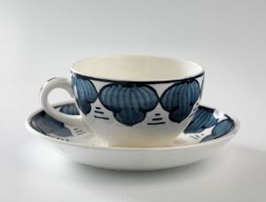  A white ceramic cup with a handle and matching saucer featuring hand-painted underglaze decorations in shades of blue, consisting of stylized leaf or scallop patterns. Designed by Andreas Schneider, the set embodies a marriage of function and art with its harmonious, rhythmic blue motifs against the glossy white background.