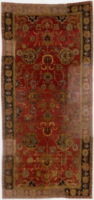  An antique rug with a rich red main field featuring symmetrical patterns in golden yellow, beige, green, navy blue, and black, with ornamental medallions and interconnected decorative elements, bordered by a lighter frame with complementary motifs.