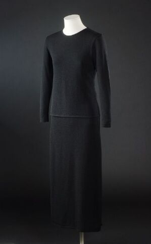  A simple, elegant black, full-length, long-sleeved dress on an off-white mannequin against a dark background, capturing a sense of timeless sophistication.