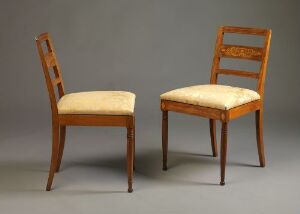  Two classic wooden chairs with vertical slat backrests and cushioned beige fabric upholstery, displayed against a plain gray background.