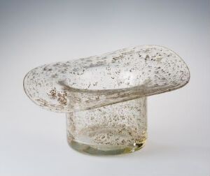 A transparent glass object with a unique mushroom-like shape, featuring a wide oval top and narrow base, with bubbles and speckles within the glass creating a dappled texture against a light grey background. Artist name and title are unknown.