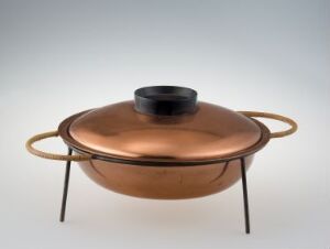  A polished copper cooking pot with a matte black lid and gold-toned handles, elevated on a black metal tripod stand against a light grey background.